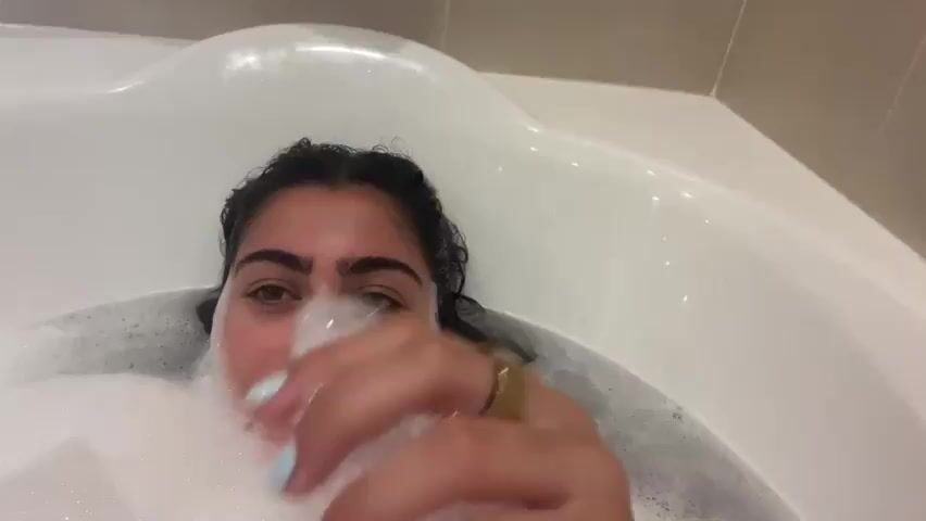A young woman with dark hair is in a bathtub filled with white foam. She is looking at the camera and smiling. Her head is mostly submerged in the foam, and she has her hand in front of her face, holding some of the foam. The bathtub is white and appears to be in a bathroom with beige tiles on the wall. The lighting is warm and somewhat dim.