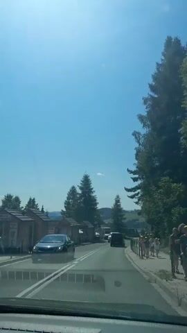 The video shows a view from inside a vehicle driving on a paved road. The road has two lanes separated by double white lines. On the left side of the road are several small wooden structures with peaked roofs, possibly market stalls or shelters. A silver car is partially visible in the lower left corner of the frame. Further down the road, a white van and a black vehicle are driving in the same direction. On the right side of the road, there is a sidewalk with a metal railing. A group of about six people, including children, are walking on the sidewalk. The environment is rural, with tall evergreen trees and hills visible in the background. The sky is clear and blue, suggesting a sunny day.