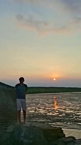 The video begins with a wide shot of a person standing on a brick structure overlooking a body of water. The sun is setting, casting a warm glow on the scene. The video then pans to show graffiti on a wall, followed by a group of people sitting and talking near several parked motorcycles. The video continues to show more parked motorcycles and people gathered by the water. The final shot shows a view of the water with trees and clouds in the background. The overall scene appears to be a public gathering spot near a body of water at sunset.