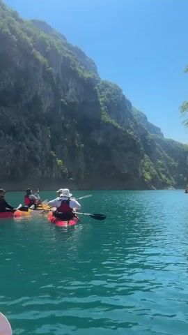 A group of four people are kayaking on a clear, turquoise lake. The kayaks are red and yellow. Two people are in one red kayak, one person is in a yellow kayak, and one person is in another red kayak. They are all wearing life vests. The kayaks are moving away from the camera and towards a large, green, mountainous cliff face. The sky is clear and blue. The water is calm. The person in the front red kayak is wearing a white hat and appears to be paddling. The person in the red kayak behind them is also paddling. The person in the yellow kayak is sitting and looking forward. The person in the back red kayak is also looking forward and appears to be paddling. The camera appears to be attached to another kayak or person in the water, as a portion of a white and pink paddle is visible in the foreground.