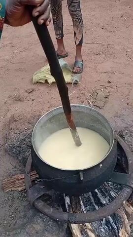 A person's hands are visible as they stir a light-colored, thick liquid in a large black pot. The pot sits over a fire with visible logs and embers. The person is holding a long, dark wooden stick with a rough texture at the stirring end. In the background, another person's legs and feet are visible, wearing dark pants with a patterned design and dark sandals. The ground appears to be dry dirt.