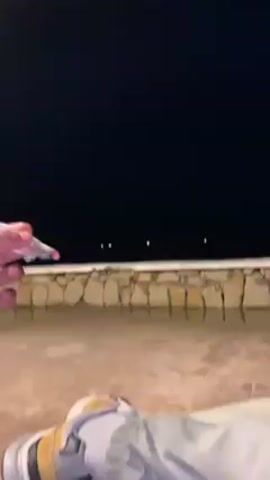 The video shows a hand holding a smoking cigarette at night. The hand is wearing a red wristband. White smoke is rising from the cigarette against the dark sky. In the background is a stone wall and what appears to be a patio.  A foot wearing a multi-colored shoe is visible at the bottom of the frame. Music plays.