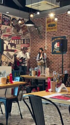 A man and a woman are performing music in a restaurant. The man is playing an electric guitar and singing. He is wearing a white shirt and dark pants. The woman is singing and playing a drum. She is wearing a light blue shirt and a dark skirt. They are standing on a small stage in front of a brick wall decorated with vintage car posters. Several tables with chairs are set up in the foreground, indicating a dining area. The lighting appears to be from overhead fixtures.