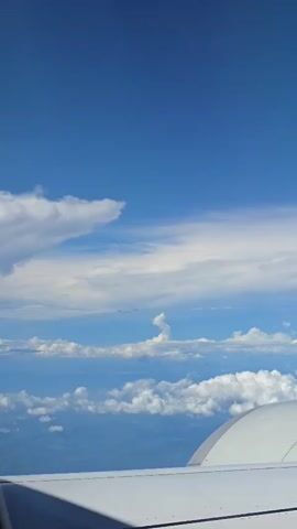 A view from an airplane window shows a vast expanse of blue sky filled with scattered white clouds. Below the clouds, a hazy landscape of mountains and possibly water is visible. The wing of the airplane, painted white, occupies the bottom right portion of the frame, with a curved edge visible against the sky. The lighting suggests it is daytime.