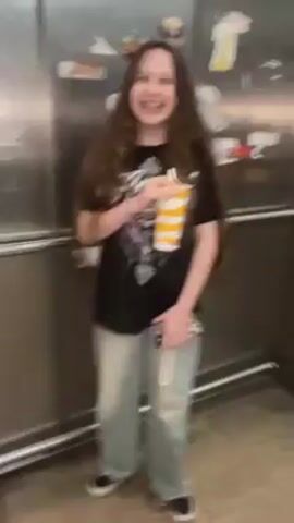 Inside an elevator, a young woman with long brown hair wearing a black t-shirt with a graphic, light blue jeans, and black and white sneakers is holding a McDonald's drink. She is laughing and shaking the drink, causing the camera to move erratically.  The elevator has stainless steel walls and a floor with beige tiles. The elevator's control panel is visible, but the buttons are blurry.