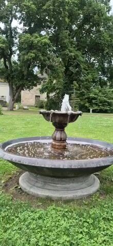 The video shows a fountain in a park or garden. The fountain is made of dark material and has water spouting from the top. The water falls into a basin, creating ripples and bubbles. The fountain is surrounded by green grass and trees. In the background, a building can be seen. The sky is overcast. The video is short and appears to be a still shot with minor movement from the water.