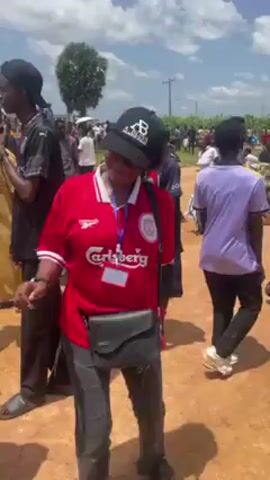 The video shows a group of people dancing outdoors on a bright, sunny day. The ground appears to be dirt or reddish soil. The person closest to the camera is wearing a red soccer jersey with white accents and a black baseball cap. They are also wearing a lanyard around their neck and have a small black purse slung across their body. This person is actively dancing, moving their arms and body to the music. Other people in the background are also dancing and socializing, creating a lively atmosphere. The crowd is diverse in terms of clothing and appearance. In the far background, trees and utility poles are visible.