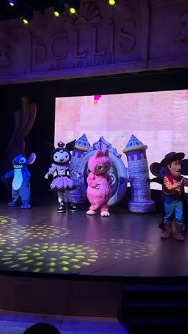 The video opens on a dimly lit stage with a large screen displaying abstract geometric patterns in shades of pink and purple. The word "BELLIS DELUXE HOTEL" is emblazoned in large, textured lettering above the screen. Five costumed characters are positioned across the stage, facing an unseen audience. On the far left is a blue character resembling Stitch from Disney. To its right is a black and white cat-like character in a pink outfit, likely Kuromi. In the center, a fluffy pink rabbit-like character stands prominently. To the right of the rabbit are two characters dressed as cowboys. The stage floor is dark and reflects yellow spotlight patterns. Two towers flank the central screen. The overall atmosphere is that of a children's performance or entertainment show.