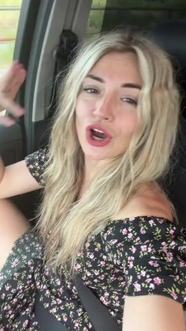 The media file features a young woman with long, blonde, wavy hair, wearing a black off-the-shoulder dress with a floral pattern, sitting in the passenger seat of a car. She is smiling and appears to be speaking or singing, with her mouth open. Her right hand is raised, and she is gesturing with her fingers. The lighting suggests it is daytime, and the background visible through the car window shows foliage and a hint of buildings, indicating an outdoor setting, possibly a rural or suburban area. The overall mood is cheerful and energetic. The visible text in the media file is "Goričani, Montenegro."