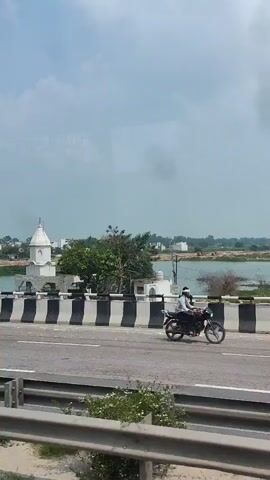 The video appears to be captured from a moving vehicle, likely a car or bus, traveling along a highway. The view is from a window, with some reflections visible. The scene showcases a white structure resembling a temple or shrine with a dome and spire, situated near a body of water. A black and white striped barrier runs along the edge of the highway, separating it from the area where the building is located. Trees and vegetation are present in the background. The sky is overcast. A dark-colored car is seen driving on the highway in the foreground. The video is short, lasting only a few seconds, and shows a landscape view with the building and water as prominent features.