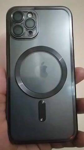 A hand holds an iPhone in a smoky-gray protective case. The back of the phone is visible. The case has a raised, round magnetic charging circle centered on the phone, around the Apple logo. There is a rectangular magnetic alignment indicator just below the circle. The camera module at the top-left corner of the case has individual covers for each of the four lenses. The background is out of focus and a cream color.