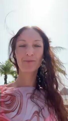 A woman is facing the camera, her face is visible and her expression is pleasant. She has long, wavy, reddish-brown hair that is blowing in the wind. She is wearing a light pink and white patterned top. Behind her is a beach with palm trees and a body of water with large rock formations in the distance. The sky is bright and clear.