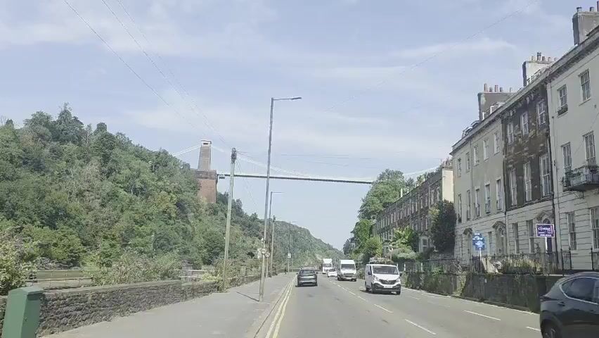 A vehicle POV shot captures a road with several cars and vans driving on it. To the left, a lush green hillside slopes downwards, with a stone wall running along the roadside and a body of water beyond. In the distance, a suspension bridge stretches across a gorge, with a tall brick tower visible on the left. To the right, a row of terraced houses with large windows and balconies lines the street. The sky is clear and blue with a few scattered clouds. The overall scene appears to be a bright, sunny day in an urban environment with natural scenery.