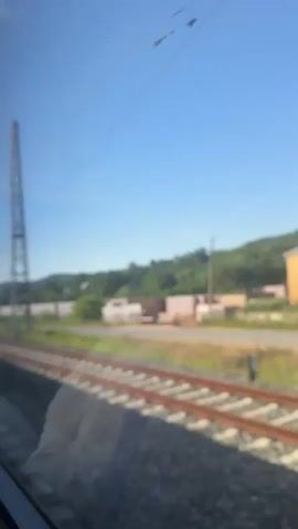 The video shows a view from a train window. In the foreground, train tracks run diagonally across the frame. A tall metal pylon with overhead wires is visible to the right. Beyond the tracks, there is a grassy area, a low wall, some trees, and a blue sky. The train appears to be moving, indicated by the motion blur. The video lasts for four seconds.