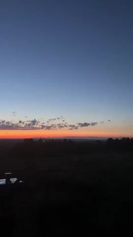 A video captures a sunset over a rural landscape. The sky transitions from a deep blue at the top to a vibrant orange and red horizon near the bottom. Silhouetted trees and hills form the dark foreground. A small, dark structure, possibly a metal post with a cap, is visible in the lower left quadrant of the frame. The scene is still and quiet, with no discernible movement or human presence.