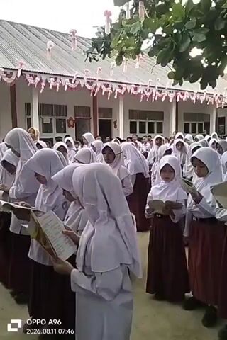 A group of female students wearing white hijabs and brown skirts are standing in front of a building. They are holding books and appear to be reading. The building has a white roof and the facade is painted white with red and white decorative banners hanging from the eaves. Trees with green leaves are visible above the students. The timestamp on the video indicates it was recorded on August 11, 2026, at 07:46. The students are arranged in rows, and the camera is positioned at their eye level, looking slightly upwards. The overall scene suggests an outdoor assembly or activity at a school.