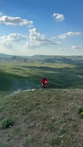 The video showcases a motocross rider on a hilltop against diverse backdrops. Initially, the rider, dressed in a red jacket and helmet, revs a red dirt bike with noticeable exhaust smoke. The backdrop is a sprawling green landscape under a blue sky dotted with fluffy clouds. The scene shifts to a woman standing with a corgi, pointing towards the horizon where the motocross bike is parked. The landscape in this part is similar, but it appears flatter. Finally, the rider is seen on the bike again, this time against a coastal backdrop with the sea visible in the distance. The sky remains blue, with some cloud cover. Another person is visible in the scene, standing off to the side.