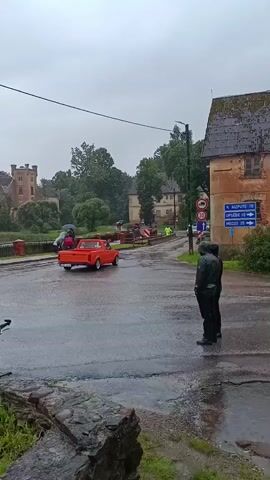 A desolate, overcast day presents a wet scene with a red vintage pickup truck driving on a wet paved road. The truck is positioned in the middle of the road, heading away from the camera. On the left side of the road, there is a bridge with a castle-like building in the background. A few people are on the bridge, some holding umbrellas. On the right side of the road, there is a person standing and facing away from the camera, dressed in dark clothing. Further down the road on the right, there is a building with a sign that has directional arrows. The ground is wet, indicating it is raining or has recently rained. The sky is cloudy and gray, suggesting a somber mood. The overall impression is one of quietness and a touch of melancholy.