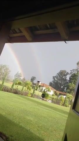 The video shows a vibrant double rainbow arching over a lush green lawn, viewed from under a covered porch.  The lawn is landscaped with palm trees and other shrubs and trees. In the background is a one-story white building with a red tile roof and a chimney. The sky is overcast, suggesting recent or ongoing rain.  The rainbow's colors are clearly visible against the gray sky. A portion of the side of a green vehicle is visible in the lower right corner of the frame.