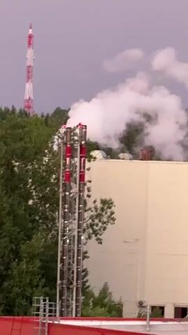 This is a vertical, handheld video. In the foreground, there is a tall, silver industrial chimney with red accents at the top. Smoke billows from the chimney. Behind the chimney is a large, cream-colored building with a red roof. In the background, there is a tall, red and white striped tower, likely a radio tower, visible above a line of green trees. The sky is overcast and a pale grey-purple color. The video is shaky, suggesting it was taken by someone holding a phone or camera.
