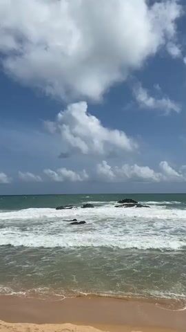 The video shows a view of a beach and the ocean on a partly cloudy day. The sand in the foreground is a light brown color. The ocean water is a greenish-blue and waves are crashing against rocks that protrude from the water. The sky is blue with scattered white clouds. The camera angle is static and facing the ocean. There are no people or animals visible in the video. The sound is of the waves crashing.