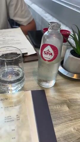 The video shows a table at a restaurant. In the foreground, there is a menu with the title "Carte des vins de l'Incontournable" which translates to "Wine list of the Unmissable." There is also a small gray bowl filled with star-shaped snacks. To the right of the snacks, there is a clear glass filled with water. Further to the right, there is a bottle of SPA brand water. In the background, a person is seated at the table, wearing a white t-shirt with text on it. There is another menu on a clipboard in front of the person. The table surface appears to be made of wood. The overall lighting suggests an indoor setting, possibly a restaurant with natural light.