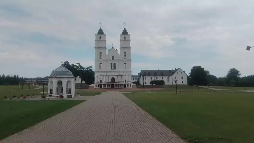 A drone shot moves forward over a paved walkway toward a white church with two spires. To the left of the church is a small white gazebo. The church is surrounded by a grassy field with trees in the background. The sky is cloudy.