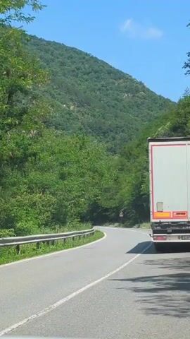The video shows a view from a moving vehicle on a winding road through a lush, green mountainous region. A large white truck with a red stripe is visible in the right lane, moving away from the camera. The road has a metal guardrail on the left side, with grass and foliage growing alongside it. The surrounding hills are densely covered with trees. The sky is clear and blue, suggesting a sunny day. A yellow traffic sign with black markings is visible on the left side of the road in the distance. The overall scene conveys a sense of travel through a scenic, natural landscape. The location is identified as Gurkovo, Bulgaria. The time of day appears to be daytime.