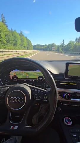 The video is filmed from inside a car, looking forward through the windshield. An Audi steering wheel, featuring the Audi rings logo and an S5 emblem, is in the foreground. The car's virtual cockpit display shows a navigation map with route guidance and a speed of approximately 105 km/h. A central infotainment screen to the right also displays navigation. The vehicle is traveling on a paved highway, with two lanes visible in its direction of travel. Green trees line both sides of the road, and the sky is clear blue. A white car is visible in the distance on the highway.