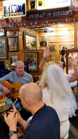 The video shows a lively scene in what appears to be a pub or bar. Several people are present, some of whom are playing musical instruments. In the foreground, a man is playing a saxophone. Behind him, a man with a gray shirt is playing an acoustic guitar and singing. To his left, another man is playing the violin. In the background, there are various decorations and advertisements, including signs for Guinness and whiskey. The overall atmosphere seems festive and communal, with people enjoying music and drinks. Some individuals are seated at tables, while others are standing or in motion. The lighting is warm and inviting, suggesting an indoor setting.