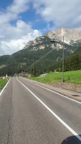 A vertical, eye-level shot captures a paved road curving slightly to the left and leading into a mountain landscape. The road has a white line down the center and a white line on the right edge. A low stone wall runs along the right side of the road, bordered by a strip of grass. Beyond the grass, a dense forest of evergreen trees covers the lower slopes of a large, rugged mountain. The mountain's peak is visible, a light brown rock face against a blue sky dotted with white clouds. Two streetlights with single lamps on tall poles are positioned along the right side of the road, with the one on the right being closer to the camera. In the distance, to the left of the road, a few buildings are visible, nestled at the base of the mountainside. The overall scene suggests a scenic, possibly remote, mountainous area.