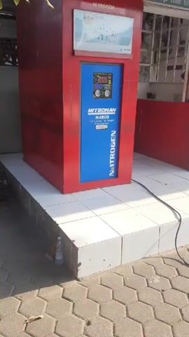 A red and blue nitrogen tire inflation machine is shown in the foreground of the video. A black cord extends from the machine and lies on a hexagonal paved surface. In the background, a person wearing a red shirt with "PERTAMINA ENDURO MOTOR SERVICE" printed on the back is attending to a white car's tire. The person is leaning over the car, suggesting they are working on the tire. A white tiled platform is situated in front of the nitrogen machine, and a clear plastic bottle is visible on the edge of the platform. The scene appears to be an outdoor service area, possibly at a gas station or auto repair shop, with a building and windows visible in the background.