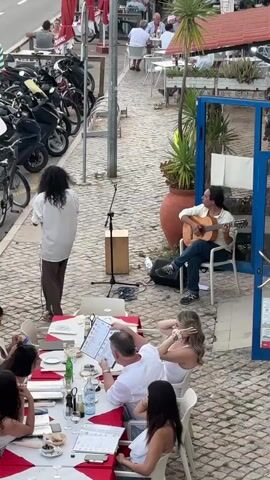 A street performer with long, dark curly hair stands at a microphone and sings, holding a microphone stand cord. To his right, another performer sits on a white chair playing an acoustic guitar. They are positioned on a cobblestone sidewalk in front of a restaurant. In the foreground, a group of people are seated at a table covered with a red and white tablecloth. Men and women are visible, some looking at menus. Behind the performers, a row of motorcycles and bicycles are parked along the street. Further in the background, other patrons are seated at outdoor tables of a restaurant. The environment appears to be a sunny day in a coastal town or city.