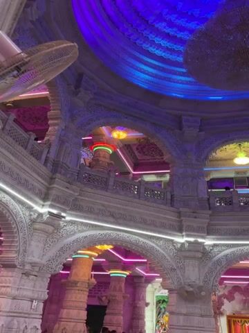 Here is a description of the video.
The video shows the interior of a building with ornate white stone architecture, illuminated with colorful LED lights. A large, round chandelier hangs from a dome-shaped ceiling adorned with a circular blue and purple pattern. Archways are visible, with some featuring colorful lighting around the pillars. People are visible on a balcony-like structure behind one of the archways. The camera is pointing upwards, emphasizing the height and detailed construction of the interior.