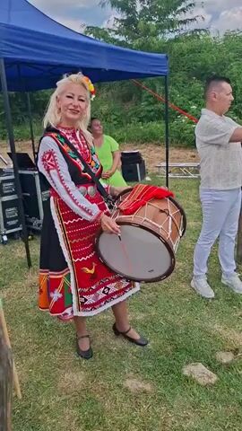 This video captures a lively outdoor scene in Shemshevo, Bulgaria, likely during a cultural event or festival. The primary subject is a woman dressed in a vibrant, traditional Bulgarian folk costume. Her attire is a rich tapestry of colors and intricate embroidery, featuring a black outer garment with red, white, and green accents, and a patterned apron. She holds a large, traditional drum with a mallet, suggesting she is about to play or is in the process of a performance. Her expression is a confident smile, hinting at engagement with the audience or her role in the event.

Beside her, a man in a light-colored, textured shirt and white pants stands with his back partially to the camera, holding up a mobile device, possibly recording the performance. In the background, another woman in a green shirt is visible, seated. The setting appears to be a grassy area with a blue tent providing shade, and some audio equipment is visible to the left, indicating a performance stage or setup. The time of day seems to be daytime, with bright lighting and a partly cloudy sky, suggesting pleasant weather. The overall atmosphere conveys a sense of celebration and cultural tradition. No visible text is present in the video.