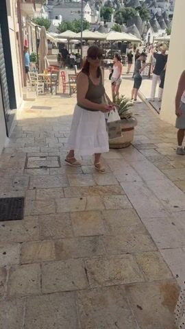 The video shows a woman walking down a cobblestone street in what appears to be a tourist town. She is wearing a dark tank top and a white skirt, and sunglasses. She is carrying a white bag over her shoulder and a pink object in her right hand. The street is lined with white buildings, some of which have balconies. In the background, there are many cone-shaped buildings with gray roofs, a distinctive architectural style. There are also outdoor tables and chairs set up in front of some of the buildings, suggesting a cafe or restaurant. Other people are visible walking in the street and sitting at tables. The sky is clear and blue.