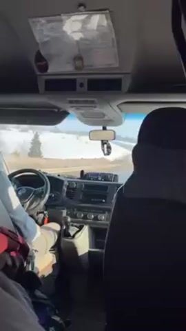 From the perspective of a rear passenger in the cab of a Volkswagen van, a fair-skinned driver in a light blue sweatshirt and white pants shifts gears. The van travels on a paved, single-lane road. Patches of snow cover the ground on either side of the road, and some evergreens dot the landscape. The interior of the van appears well-used. A piece of paper is attached above the rearview mirror, and a black pillow rests on the headrest of the seat in front of the camera.