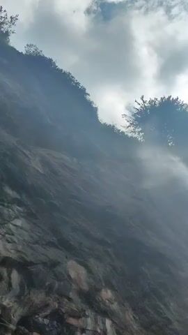 This media file depicts a dramatic natural scene in Solang, India. The primary subject is a sheer rock face, steeply inclined and textured, suggesting a cliff or mountainside. Lush green foliage, including trees and bushes, clings to the upper edge of the rock face, creating a stark contrast between the rugged stone and the vibrant life it supports.

A significant element of the scene is the presence of mist or spray, possibly from a waterfall or heavy condensation, obscuring parts of the rock and creating a hazy, ethereal atmosphere. This mist catches the light, giving it a luminous quality. Above the rock face, the sky is visible, filled with a dynamic arrangement of clouds. Some are bright and white, illuminated by sunlight, while others are darker and more diffused.

The lighting suggests it is daytime, with the sun breaking through the clouds, casting rays that create lens flares and add to the mystical ambiance. The sun's position, partially obscured by trees and clouds at the top right, hints at either early morning or late afternoon. The overall mood is one of awe and natural grandeur, with the mist adding an element of mystery. There are no discernible people or man-made objects in the frame, emphasizing the raw, untamed beauty of the natural environment. The location context of Solang, India, is a well-known mountainous region, which aligns with the visual cues of steep terrain and lush vegetation.