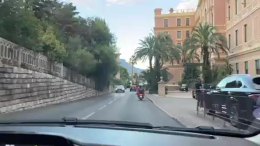 From the inside of a moving vehicle, a driver's perspective is revealed on a street flanked by trees and stone walls on the left, and a large building with palm trees on the right. Cars and motorcycles are on the road. A man stands on the sidewalk on the right. Music is playing.