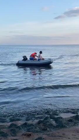 Two people are in a small inflatable boat on the water. The person in the front is wearing a life jacket and appears to be steering the boat with a tiller attached to an outboard motor. The person in the back is wearing a white shirt and dark shorts. The boat is moving away from the shore, where a small amount of dark-colored seaweed or debris is visible on the sand. The water is calm, with gentle ripples. The sky is a pale blue with some light clouds. The overall scene suggests a leisurely boat ride on a calm day.