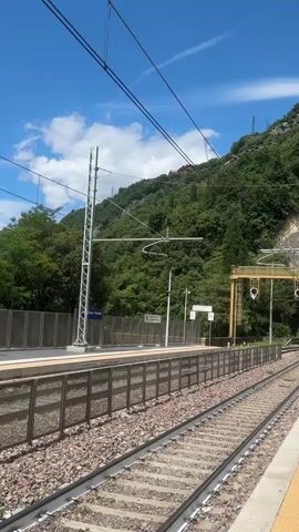 The video begins by showing a train station platform with a rocky, tree-covered mountain slope in the background. The sky is blue with some clouds. Train tracks run along the platform, and overhead electrical wires are visible. As the video progresses, the camera pans to the right, revealing a train tunnel entrance in the mountain. A person in dark clothing stands on the platform near the tunnel. The camera continues to pan right, showing more of the platform and the mountain. A blue sign with white lettering that reads "Varenna - Es" is visible at the end of the video. The video appears to have been filmed on a sunny day.
