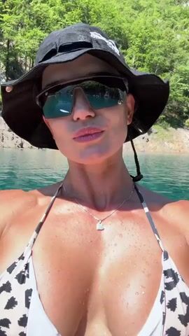 A woman is in a kayak on a body of water. She is wearing a black bucket hat, sunglasses, and a patterned bikini top. She is smiling and looking at the camera. The water is a clear blue-green color, and the background shows a forested hillside. The kayak is yellow and white. The video is a selfie, taken from the woman's perspective.