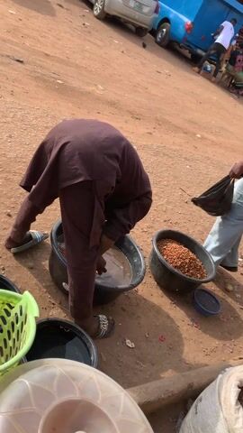 The scene is a dusty, outdoor marketplace, with several individuals present. In the foreground, a person dressed in a brown outfit is bent over, washing what appears to be peanuts in a large black tub filled with murky water. Another tub filled with dry peanuts sits beside it. To the left, there's a green basket and a clear plastic container. In the background, a woman in a white shirt and light jeans stands with a black bag in her hand, looking to her right. Another individual in a golden outfit walks by, facing away from the camera. In the far background, there's a blue van, other individuals, and market stalls. The ground is covered in reddish-brown dirt and scattered debris.