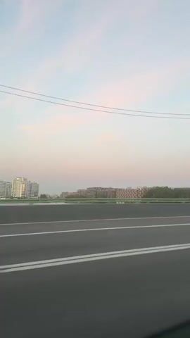 This video captures a daytime view from a moving vehicle, likely a car or bus, on a highway or elevated road in Saint Petersburg, Russia. The scene unfolds against a soft, pastel sky painted with wispy clouds in shades of pale blue and pink, suggesting either sunrise or sunset.

The foreground is dominated by the asphalt road with clear white lane markings, indicating a well-maintained thoroughfare. A green barrier separates the road from the landscape beyond. In the middle ground, a body of water, possibly a river or canal, is visible.

Across the water, a city skyline emerges. Several modern buildings with white and light-colored facades are prominent, reflecting the gentle light. Further into the distance, other buildings and lush green trees create a layered urban and natural landscape. A tall, slender lamppost is a notable object in the scene, appearing in the middle of the frame for a brief period, its structure blurred by motion.

During the clip, a bright yellow bus passes by in the opposite direction, its windows dark and indistinct, a common sight on urban transport routes. The motion of the vehicle carrying the camera creates a sense of travel and forward momentum, with the scenery subtly shifting and blurring.

The overall mood is serene and tranquil, evoked by the soft lighting and the expansive sky. The urban elements are softened by the natural light and the presence of greenery, creating a peaceful impression of a city at the edge of day. No visible text is present in the video.