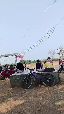 The video depicts an outdoor scene on a sunny day with a clear sky. Several people are present, some sitting and others standing. In the foreground, a blue mountain bike is parked in front of a low concrete structure. A few motorcycles are visible in the background. Long, thin objects, possibly kites or streamers, are suspended in the sky, creating a decorative effect. A white and red flag is also seen attached to a pole. The ground is covered with dry grass and dirt. The overall atmosphere appears casual and social, possibly indicating an outdoor event or gathering.