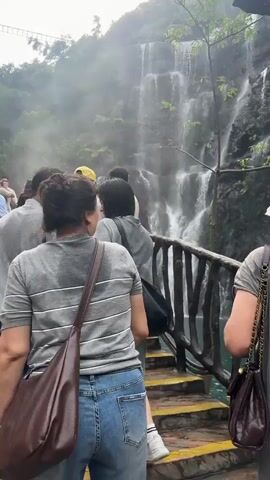 A group of people are walking along a wooden railing in front of a multi-tiered waterfall. The waterfall is cascading down a rocky cliff face, with mist rising from its base. The people are casually dressed, and some are looking at the waterfall, while others are talking amongst themselves. The environment is lush and green, with trees and foliage growing on the rocks.
