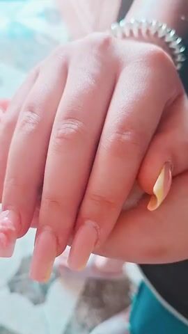 The video opens with a close-up of a person's hands, displaying long, light-pink acrylic nails. The hands are cupped together, and two small, light-brown and white striped hamsters are nestled within them. The hamsters have red eyes and visible white ear tufts. The background is slightly blurred, suggesting a shallow depth of field, with hints of pastel colors and a glimpse of a beaded bracelet on the person's wrist. The lighting is soft and diffused.