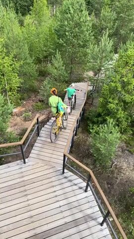 Two people, a woman and a child, are walking up a wooden boardwalk with bicycles. The woman is in front, wearing a green shirt and yellow headscarf, and is pushing a yellow bicycle. The child is behind her, wearing a green shirt and white cap, and is pushing a white bicycle. The boardwalk is surrounded by trees and vegetation, suggesting an outdoor, possibly wooded, environment. The boardwalk has wooden planks and black metal railings. The camera is positioned above and behind the individuals, looking down at them as they ascend the boardwalk.