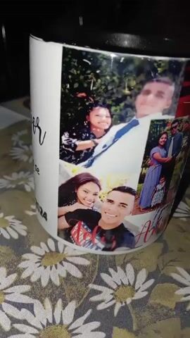 Here's a detailed description of the video:

The video shows a white mug with a collage of photos printed on it. The mug has a black interior and rim. The photos appear to depict a young couple in various settings, including formal events and casual selfies. Some photos also include other people, possibly family members, including a child. The photos are arranged in a collage style, covering the surface of the mug.

The mug sits on a tablecloth with a floral pattern of white daisies with yellow centers. The camera slowly pans around the mug, showing the different photos in the collage. The overall impression is of a personalized gift item. The words “Amor” and the date “2024” are visible on the mug.