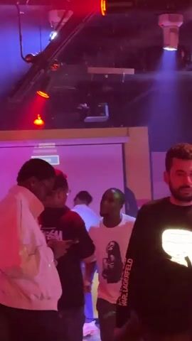The video shows a nightclub scene with people socializing and dancing. There are groups of people standing near a projector screen, and others are scattered throughout the room. Some individuals are wearing casual clothing, while others appear more dressed up. The lighting is colorful and dynamic, with red, blue, and green hues dominating the space. There are tables with glasses and cans of beer visible in the foreground, and the ceiling has exposed ductwork and lighting fixtures. The overall atmosphere is lively and energetic.